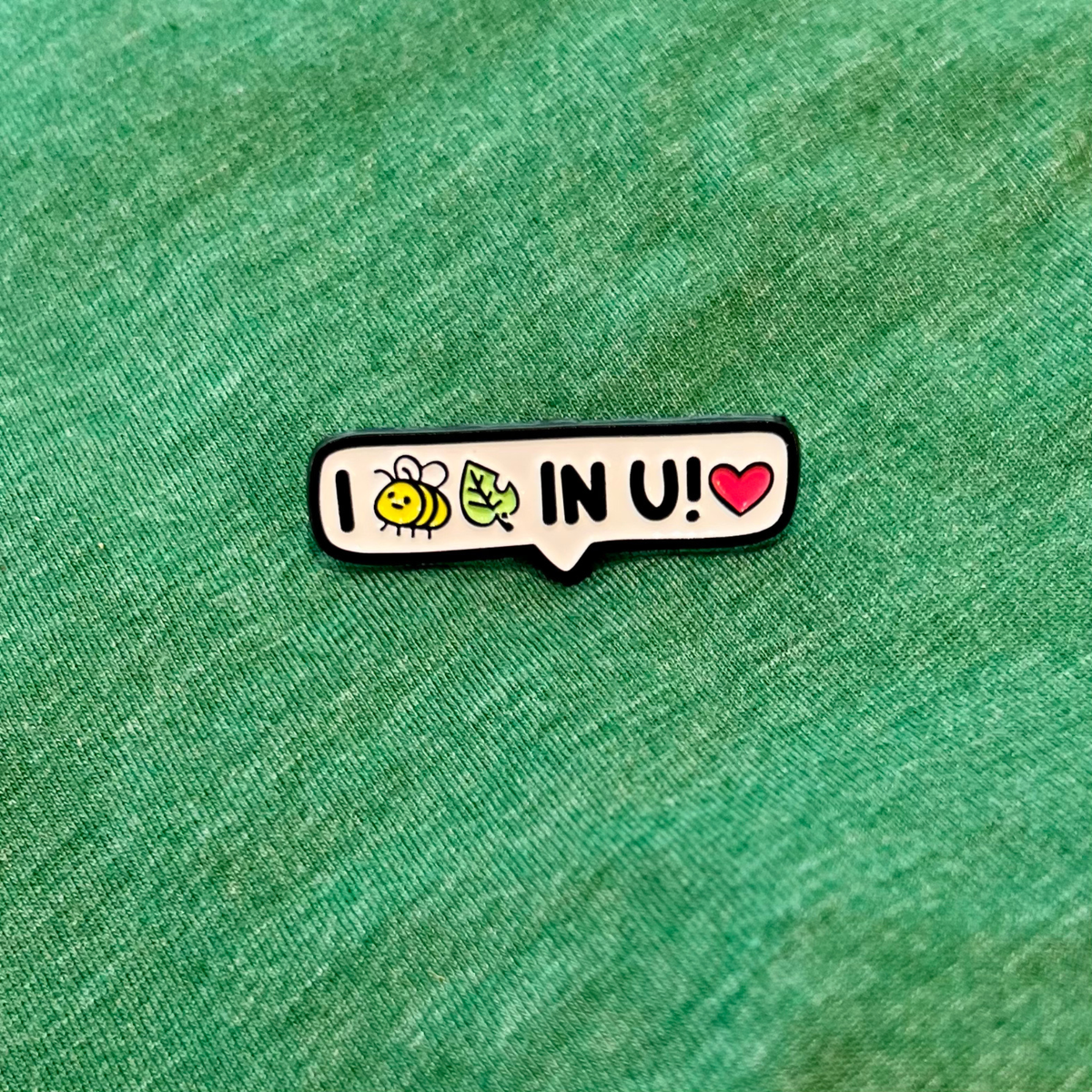 A small enamel pin shaped like a speech bubble with the text 'I (bee icon and leaf icon - read as BELIEVE) IN U!' and cute icons, placed on a textured green fabric background.