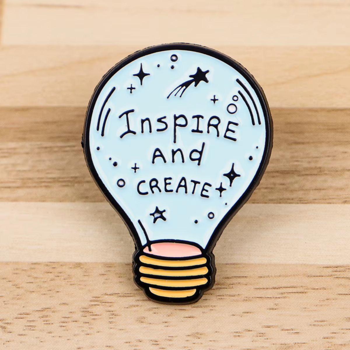 A lightbulb-shaped enamel pin with the words 'Inspire and Create' in black lettering on a pale blue background, with black stars and dots, a small shooting star, and a yellow-to-pink striped screw base, photographed on a light wood surface.
