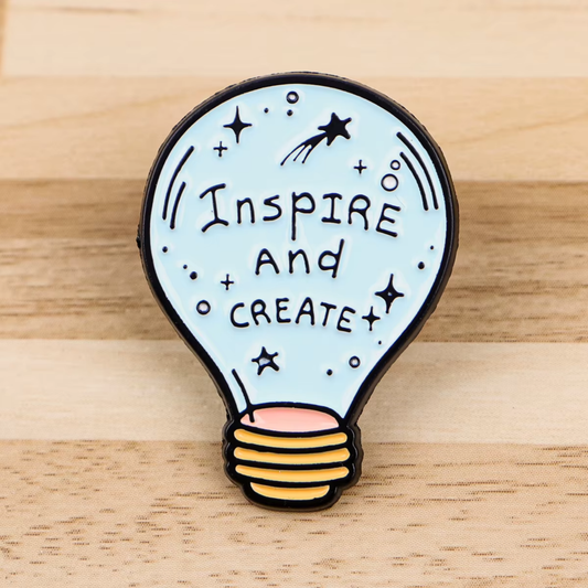 A lightbulb-shaped enamel pin with the words 'Inspire and Create' in black lettering on a pale blue background, with black stars and dots, a small shooting star, and a yellow-to-pink striped screw base, photographed on a light wood surface.