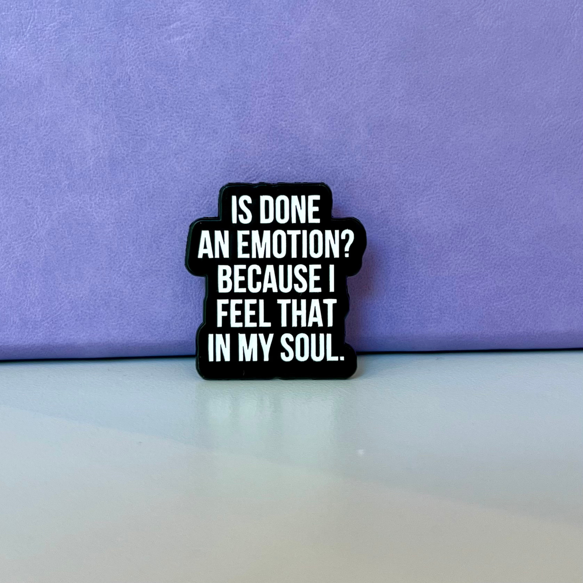 A small black enamel pin with bold white text reading 'IS DONE AN EMOTION? BECAUSE I FEEL THAT IN MY SOUL.' displayed on a light surface against a textured purple background.