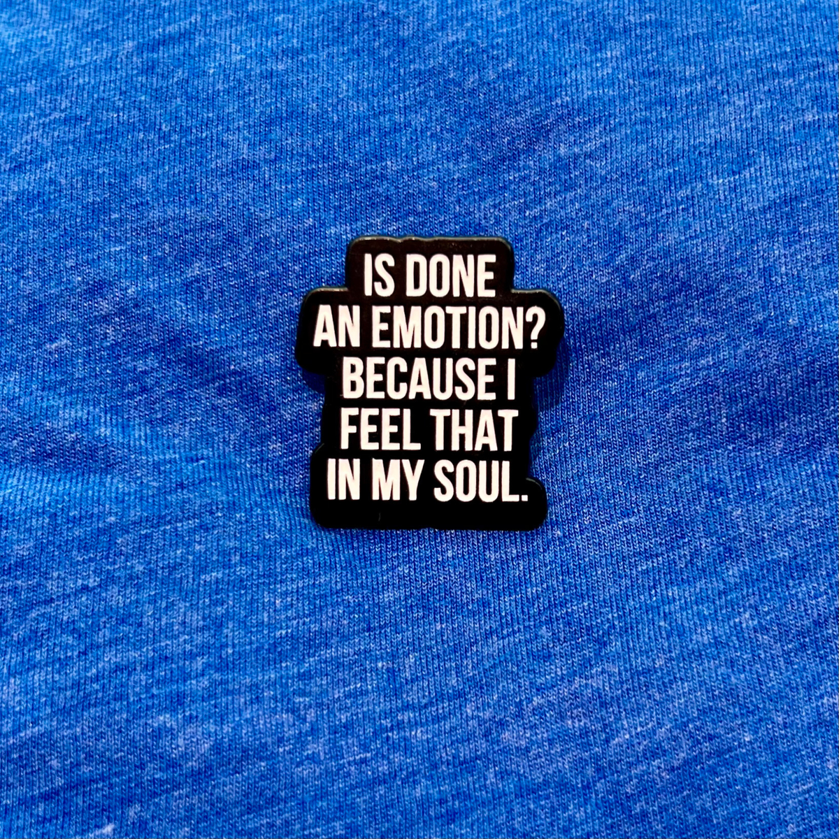 A black enamel pin with white text reading 'IS DONE AN EMOTION? BECAUSE I FEEL THAT IN MY SOUL.' displayed on a textured blue fabric background.