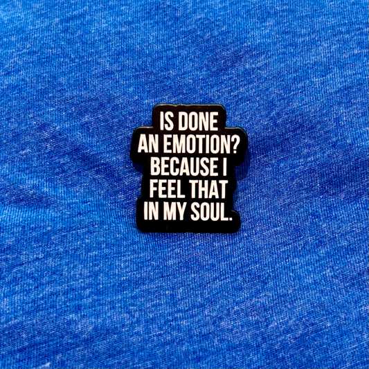 A black enamel pin with white text reading 'IS DONE AN EMOTION? BECAUSE I FEEL THAT IN MY SOUL.' displayed on a textured blue fabric background.