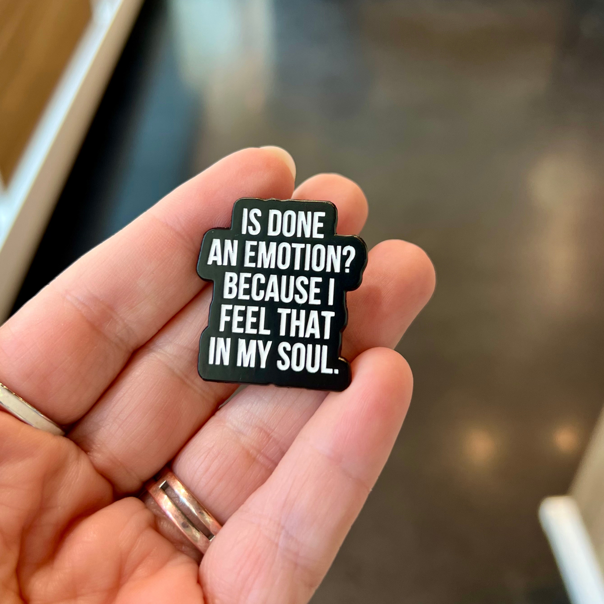 A hand holding a small black enamel pin with white text that reads, 'IS DONE AN EMOTION? BECAUSE I FEEL THAT IN MY SOUL.'