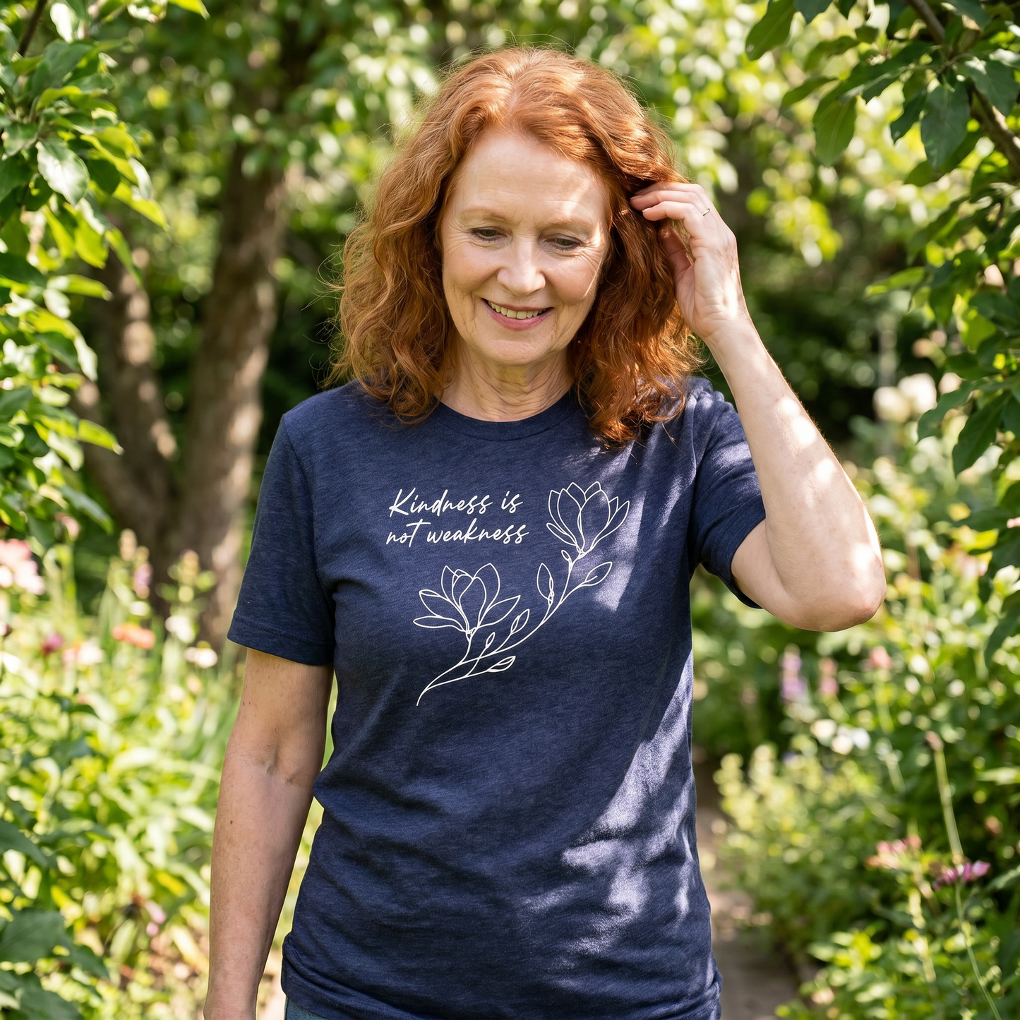 A smiling older woman with red hair strolls through a lush garden, wearing the “Kindness is Not Weakness” T-shirt by RainBee Designs LLC, featuring inspirational florals and an uplifting message.