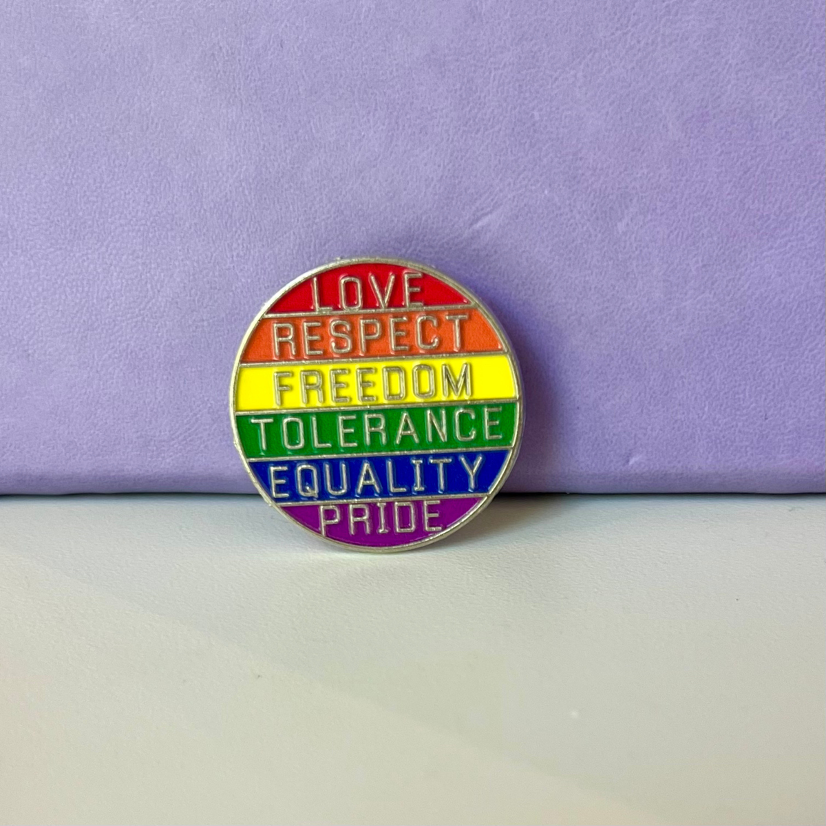 A circular rainbow pride enamel pin with silver lettering reading 'LOVE RESPECT FREEDOM TOLERANCE EQUALITY PRIDE' on a light gray surface against a lavender background.