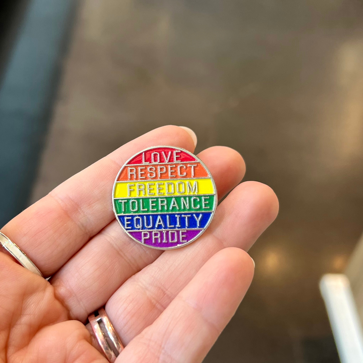 A hand holding a circular enamel pin with the words 'LOVE RESPECT FREEDOM TOLERANCE EQUALITY PRIDE' in rainbow stripes.