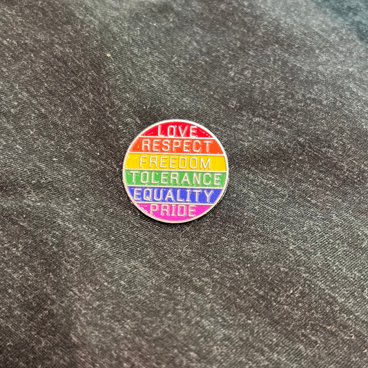 A circular enamel pin with the words 'LOVE RESPECT FREEDOM TOLERANCE EQUALITY PRIDE' in white text over rainbow stripes, placed on dark gray fabric.
