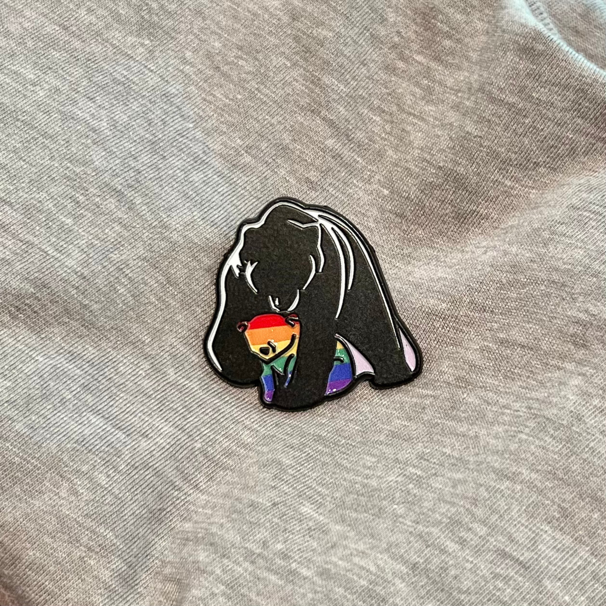 A close-up of a black enamel pin featuring a bear silhouette holding a rainbow-striped smaller bear, placed on a heather gray fabric background.