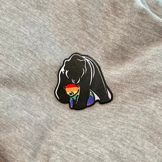 A close-up of a black enamel pin featuring a bear silhouette holding a rainbow-striped smaller bear, placed on a heather gray fabric background.