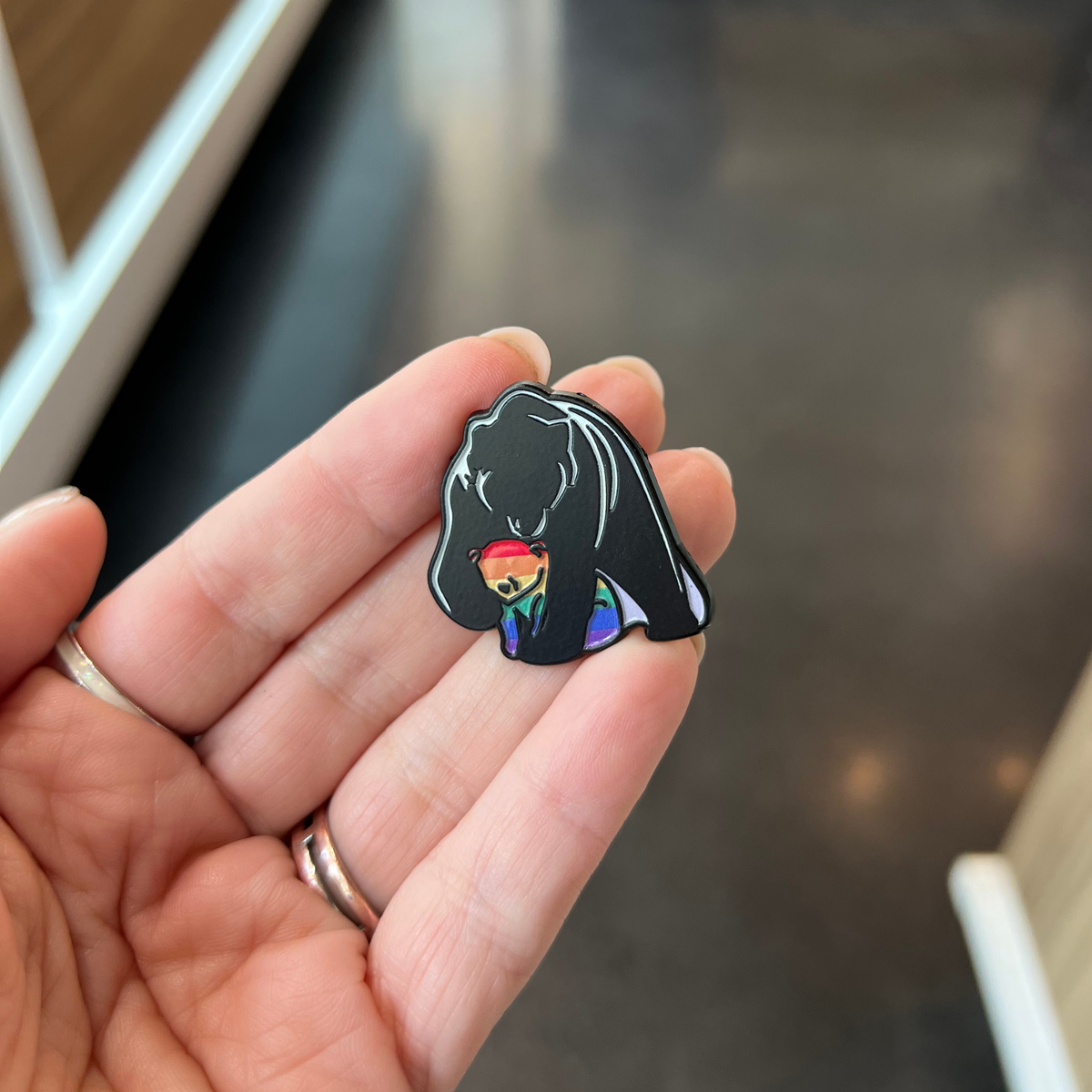 A hand holding a small enamel pin shaped like a black bear holding a smaller rainbow-striped bear, with a blurred indoor background.
