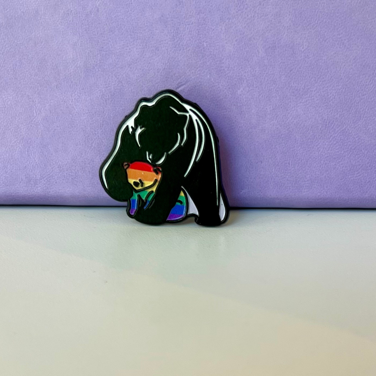 A black bear-shaped enamel pin with white line details holding a small rainbow bear, photographed on a white surface against a textured purple background.