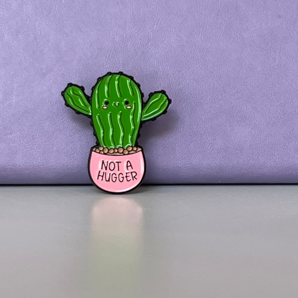 A small enamel pin shaped like a green cactus in a pink pot, with the text 'NOT A HUGGER' on the pot, photographed against a lavender background and light gray surface.