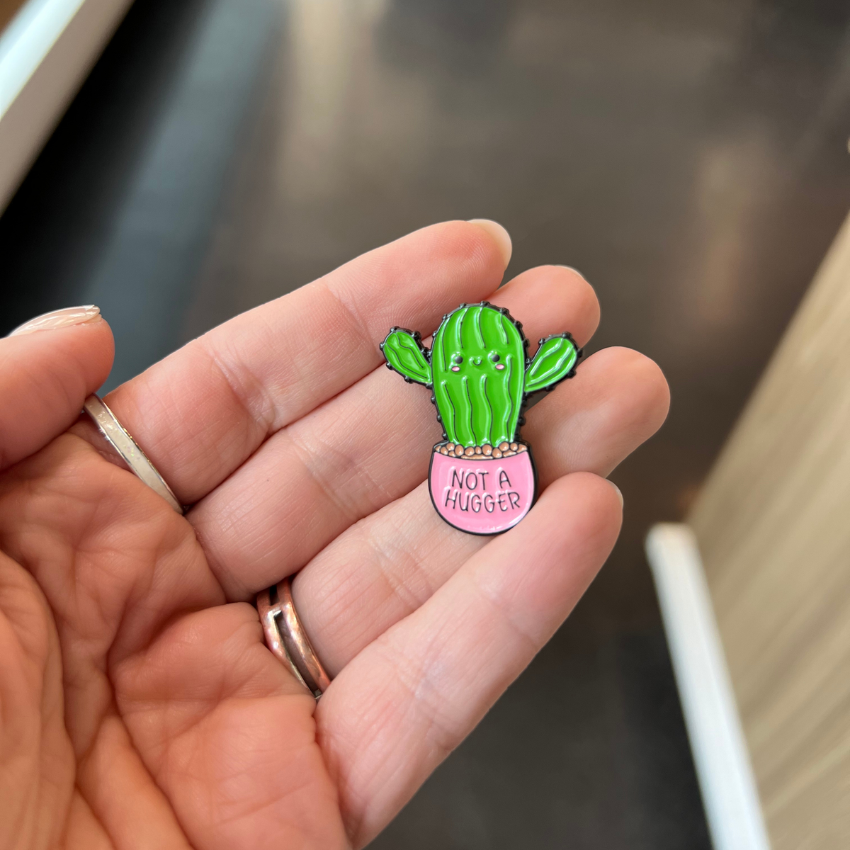 A hand holding a small enamel pin shaped like a green cactus in a pink pot with the text 'NOT A HUGGER'.