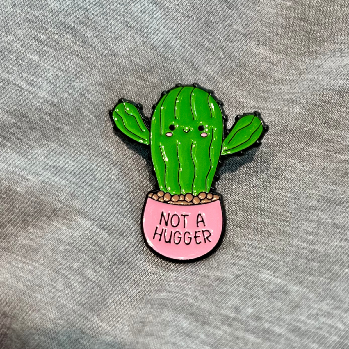 A cute enamel pin of a green cactus in a pink pot with the text 'NOT A HUGGER' on a gray textured fabric background.