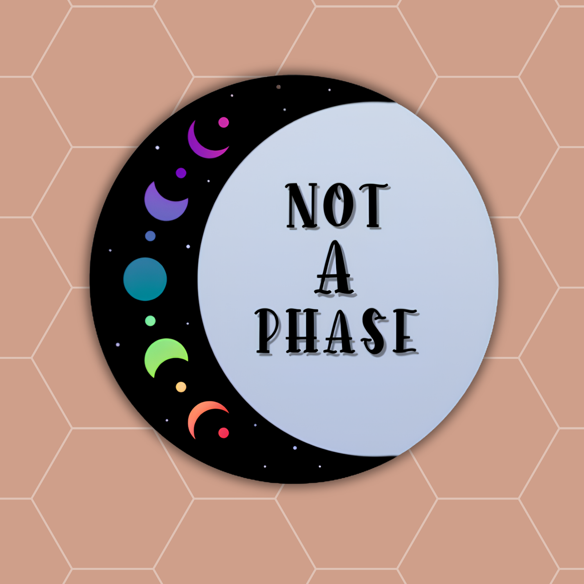 A circular sticker or decal featuring a crescent moon design with the text 'NOT A PHASE' in black lettering on a pale blue section, with a black crescent side decorated with colorful moon phases. The sticker is centered on a peach background with a hexagonal grid pattern.