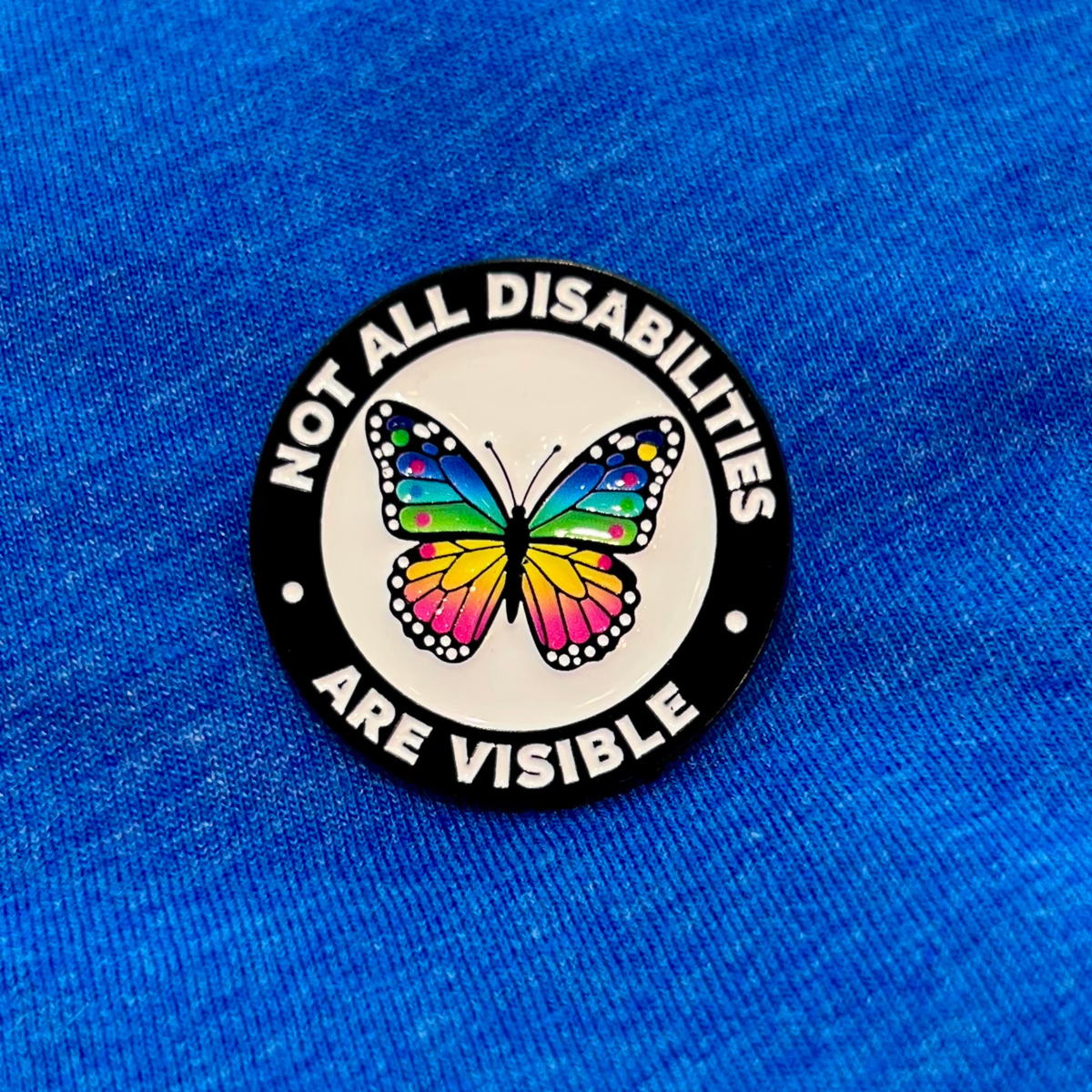 A circular enamel pin with a black border and white center, featuring a colorful butterfly and the text 'NOT ALL DISABILITIES ARE VISIBLE' in white, placed on a textured blue fabric background.