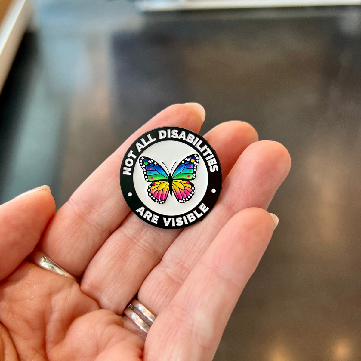 A close-up of a hand holding a round enamel pin that reads 'NOT ALL DISABILITIES ARE VISIBLE' around a colorful butterfly on a black and white background.