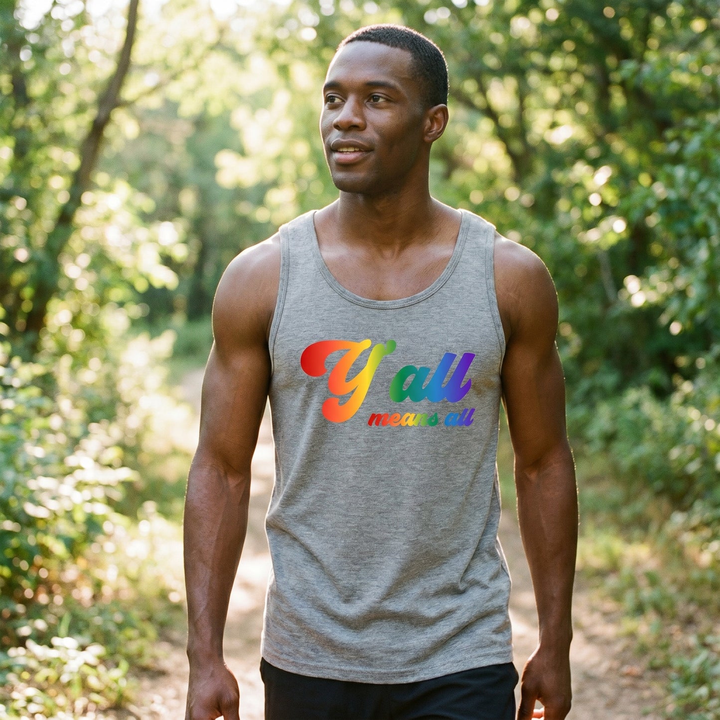 A man walks along a sunlit forest trail wearing the Y'all Means All Top by RainBee Designs LLC from the Pride Collection, its rainbow text standing out amidst the trees and greenery—an unmistakable statement of inclusivity.