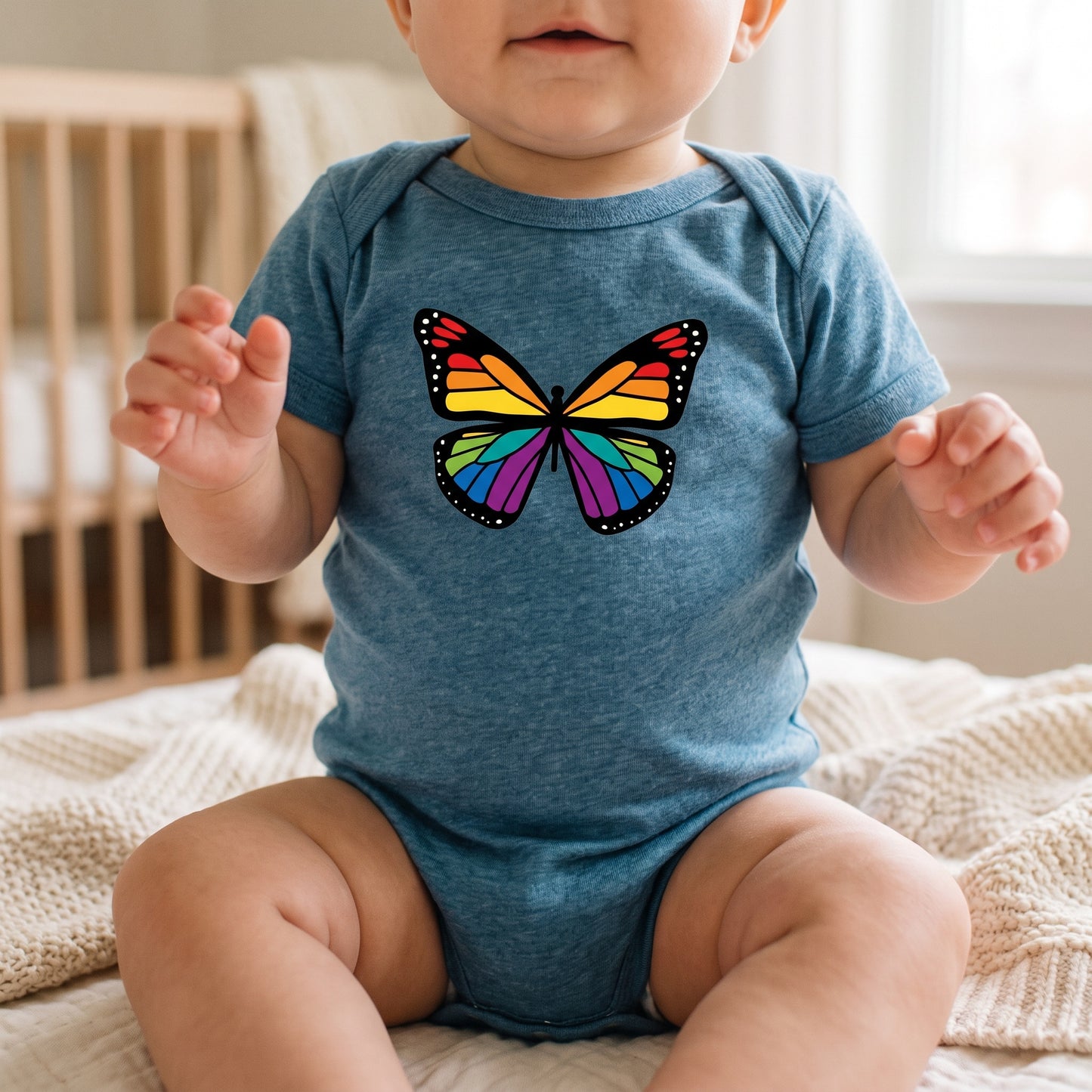A baby wearing a cozy RainBee Designs LLC Rainbow Butterfly Infant Onesie, featuring a colorful butterfly graphic, sits on a soft blanket in a gently lit room with a crib in the background.