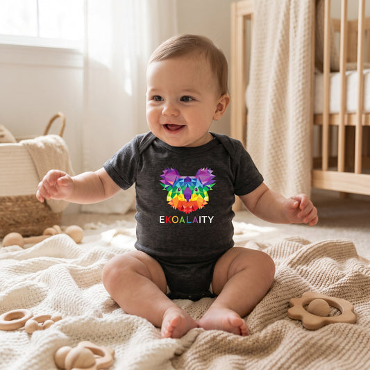 A smiling baby sits on a blanket in a cozy room, wearing the EKOALAITY Infant Onesie by RainBee Designs LLC. The dark onesie features a colorful geometric koala in pride colors with "EKOALAITY" printed on it, while wooden toys are scattered around.