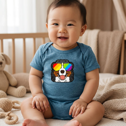 A smiling baby in a blue RainBee Designs LLC Bernese Mountain Dog (Berner) Infant Onesie, featuring a cartoon pup with rainbow sunglasses, sits on a bed in a cozy, softly lit nursery with a teddy bear and blankets in the background.