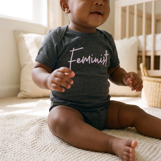 A baby sits on a soft carpet in a sunlit room, wearing the RainBee Designs LLC Feminist Infant Onesie in dark gray, featuring "Feminist" written in cursive across the chest. This adorable statement-making outfit is perfect for your little activist. A crib and basket can be seen in the background.