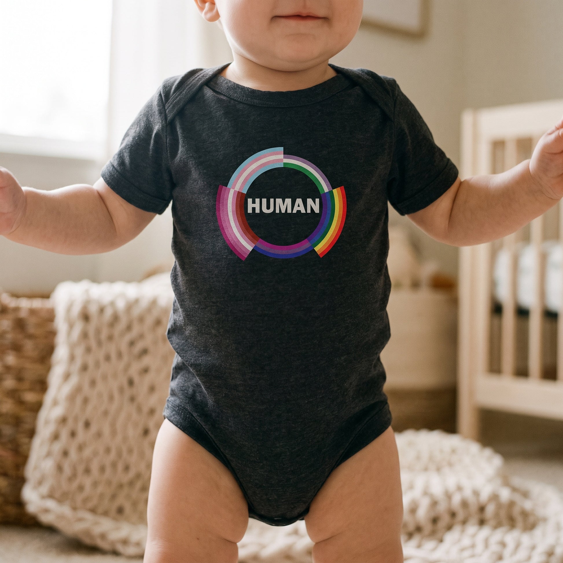 A baby stands wearing the "Human Infant Onesie" by RainBee Designs LLC, crafted from soft cotton and adorned with a vibrant pride-colored circle and the word HUMAN on the front. In the softly lit background, a crib and knitted blanket can be seen.