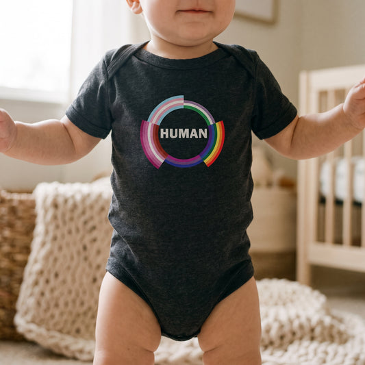 A baby stands wearing the "Human Infant Onesie" by RainBee Designs LLC, crafted from soft cotton and adorned with a vibrant pride-colored circle and the word HUMAN on the front. In the softly lit background, a crib and knitted blanket can be seen.