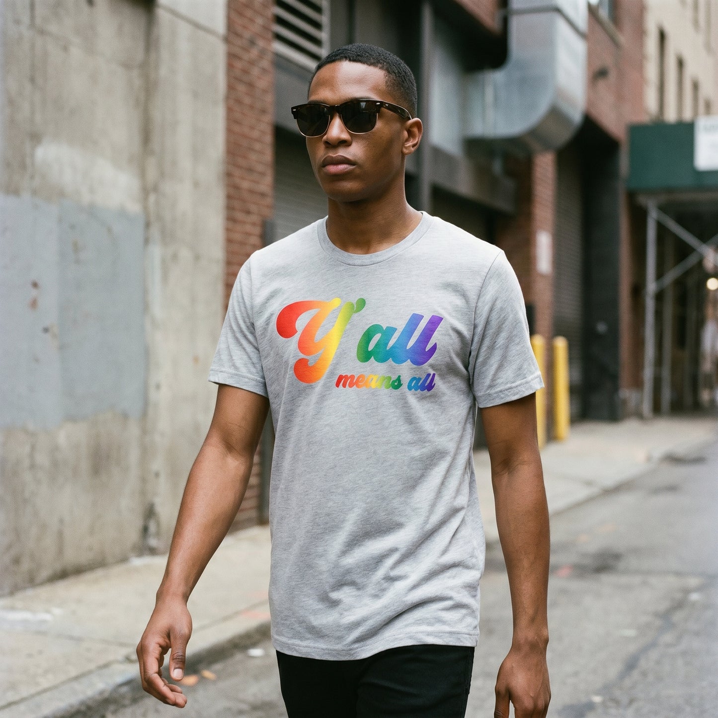 Wearing sunglasses and a gray "Y'all Means All Top" from RainBee Designs LLC, part of the Pride Collection, a person walks down a city street lined with brick buildings, celebrating inclusive apparel.