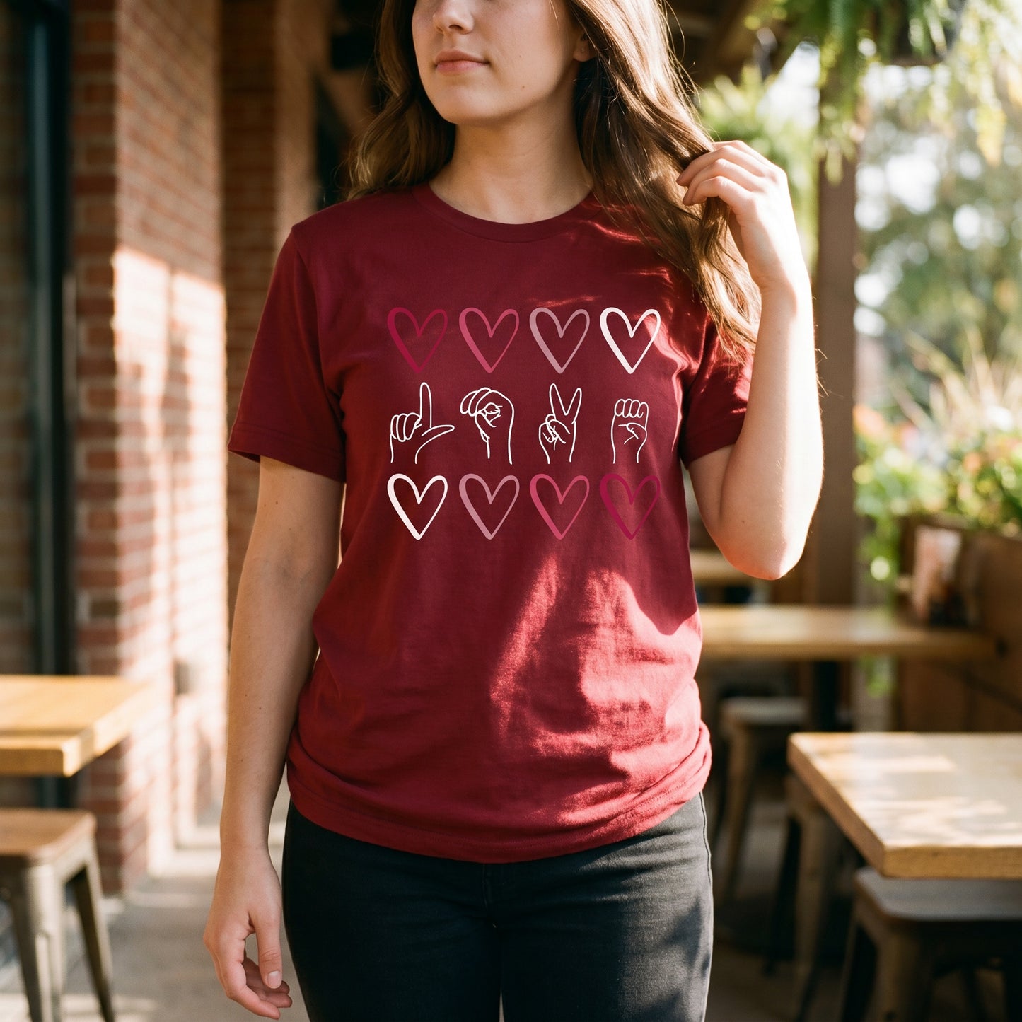 A woman wears the RainBee Designs LLC Sign Language Shirt, a cardinal red tri-blend tee featuring hearts and American Sign Language hand signs on the front, as she stands outdoors near tables.