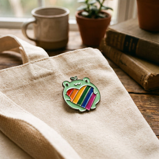 A beige tote bag adorned with a charming "Frog holding Pride Heart" pin by RainBee Designs LLC and an LGBTQ enamel pin rests on a wooden surface beside stacked books, potted plants, and a mug by the window.