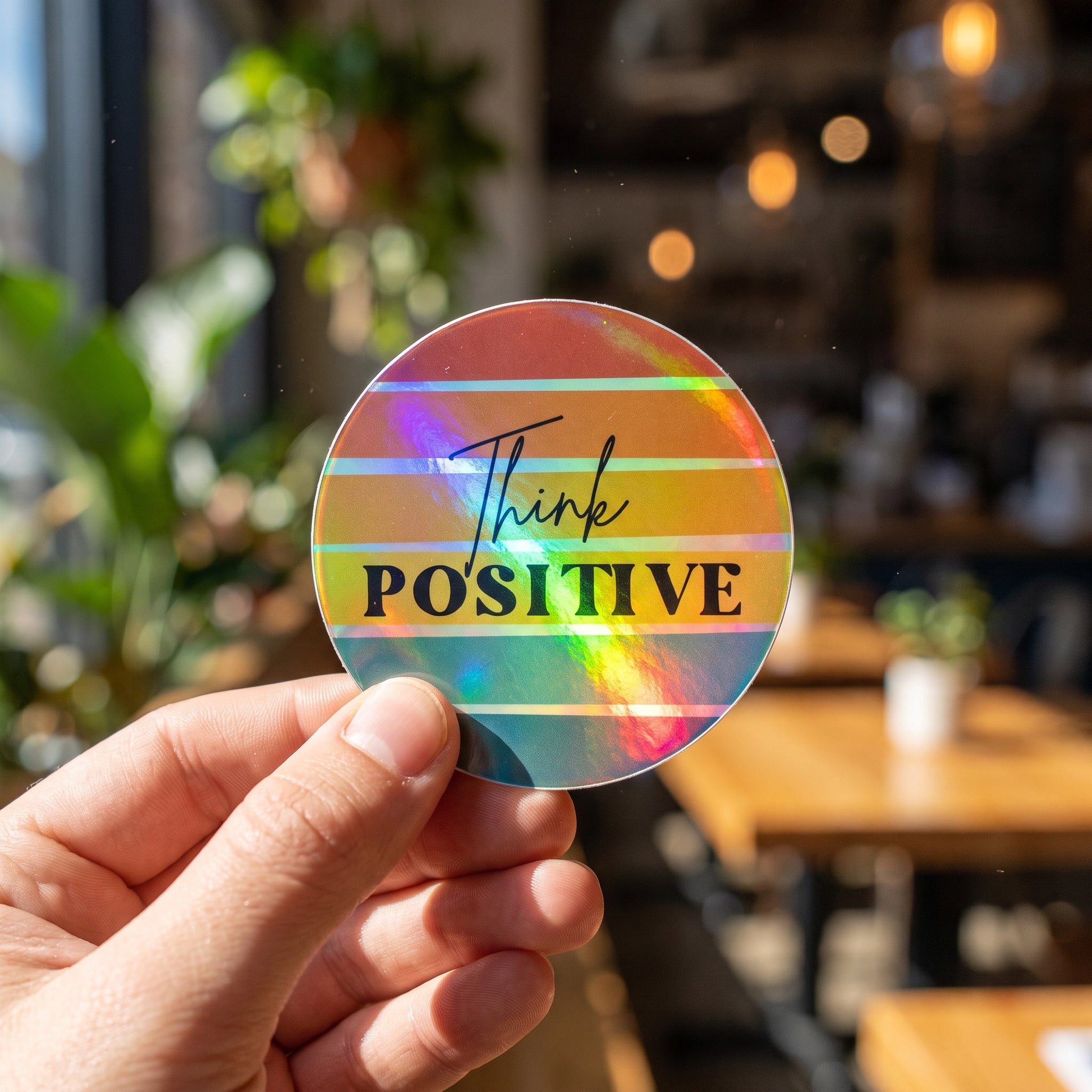 A hand holds a round, holographic Think Positive Sticker by RainBee Designs LLC, featuring rainbow stripes, in a bright, sunlit cafe filled with plants and wooden tables in the background.