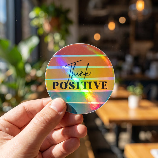 A hand holds a round, holographic Think Positive Sticker by RainBee Designs LLC, featuring rainbow stripes, in a bright, sunlit cafe filled with plants and wooden tables in the background.