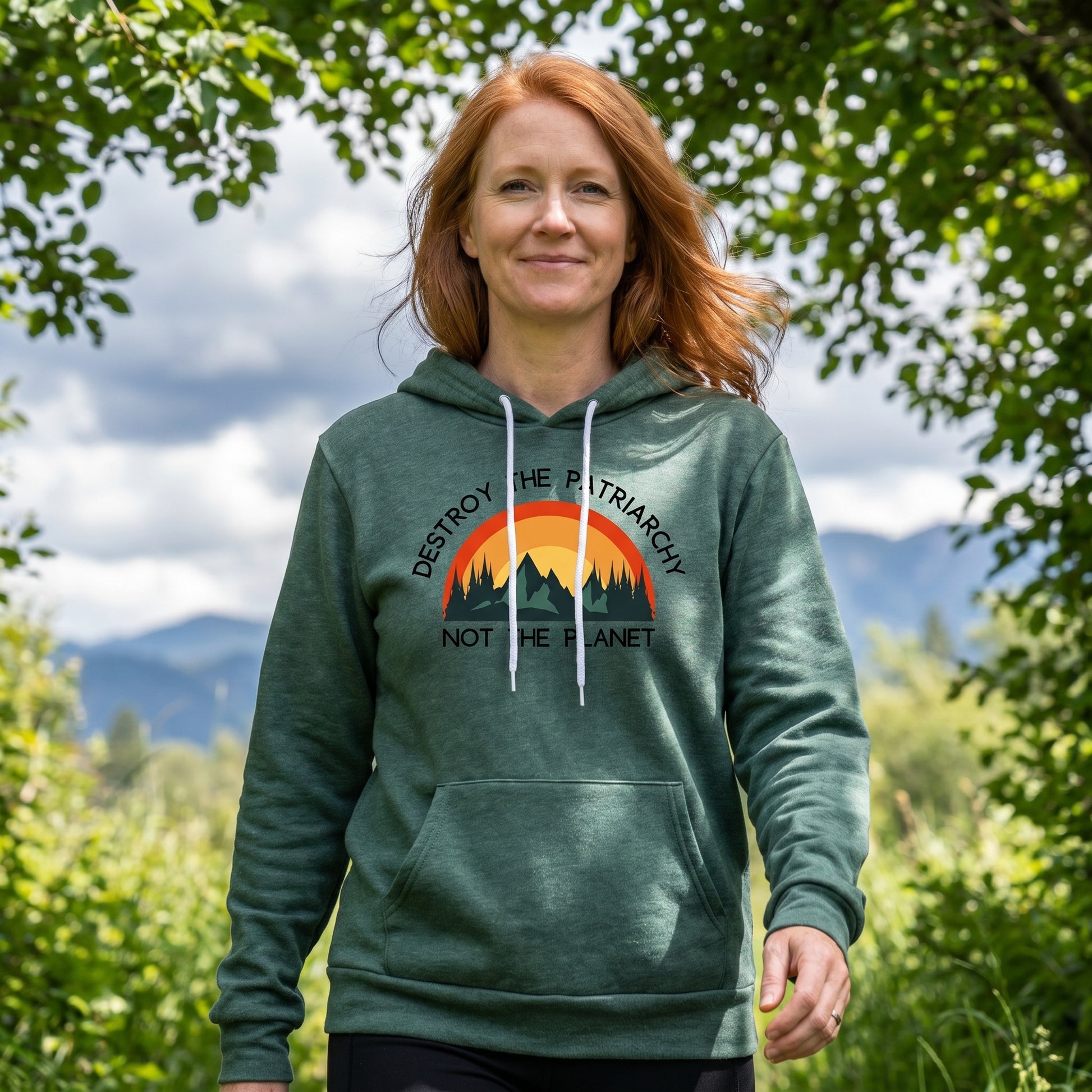 A woman with long red hair walks outdoors, smiling. She wears the Green Destroy the Patriarchy Not the Planet Hoodie by RainBee Designs LLC, which features a sunset mountain graphic and the words "Destroy the Patriarchy, Not the Planet." Trees and mountains are in the background.