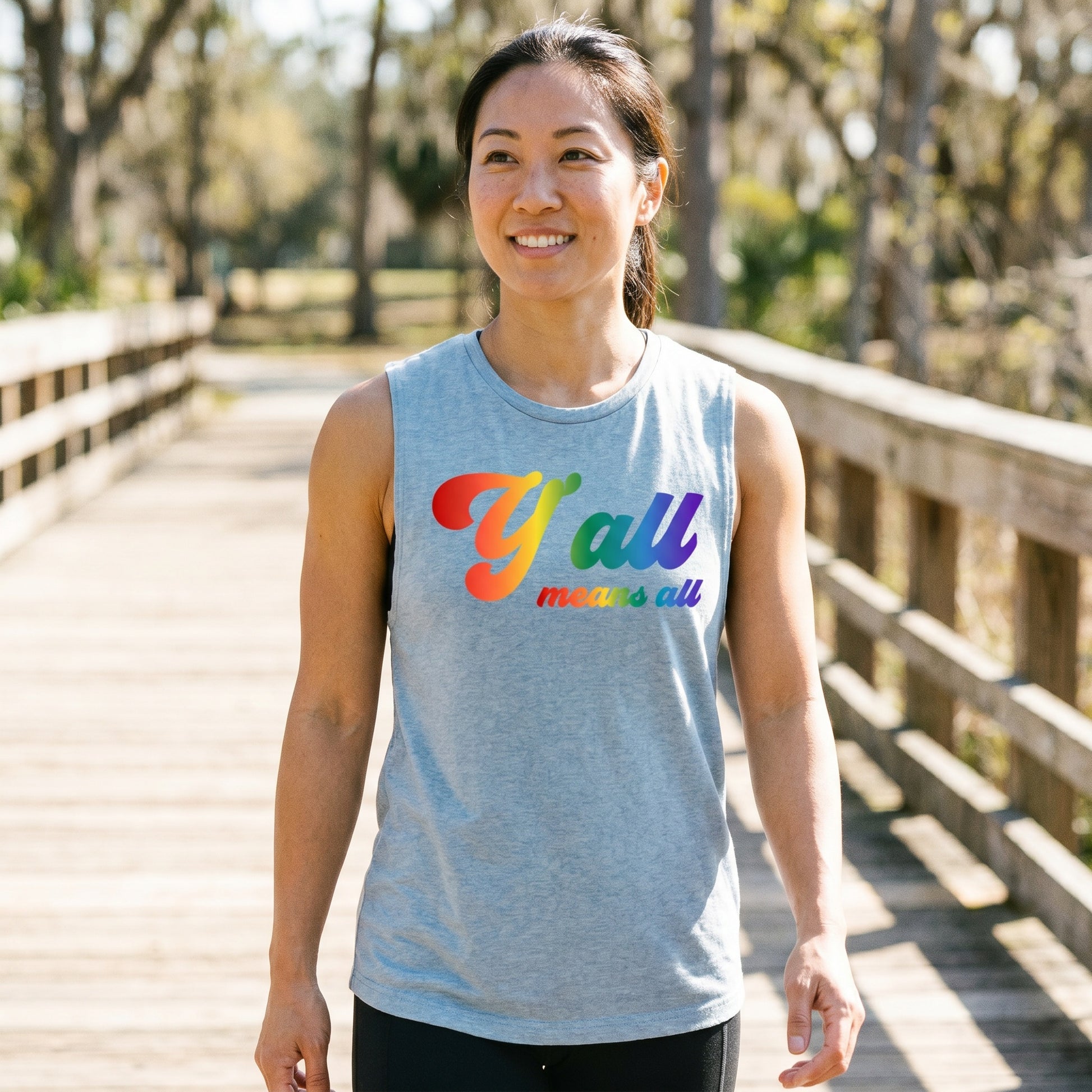 A person smiles while walking on a wooden bridge, wearing the Y'all Means All Top by RainBee Designs LLC from the Pride Collection—a gray sleeveless shirt featuring rainbow text. Trees and sunlight are visible in the background.