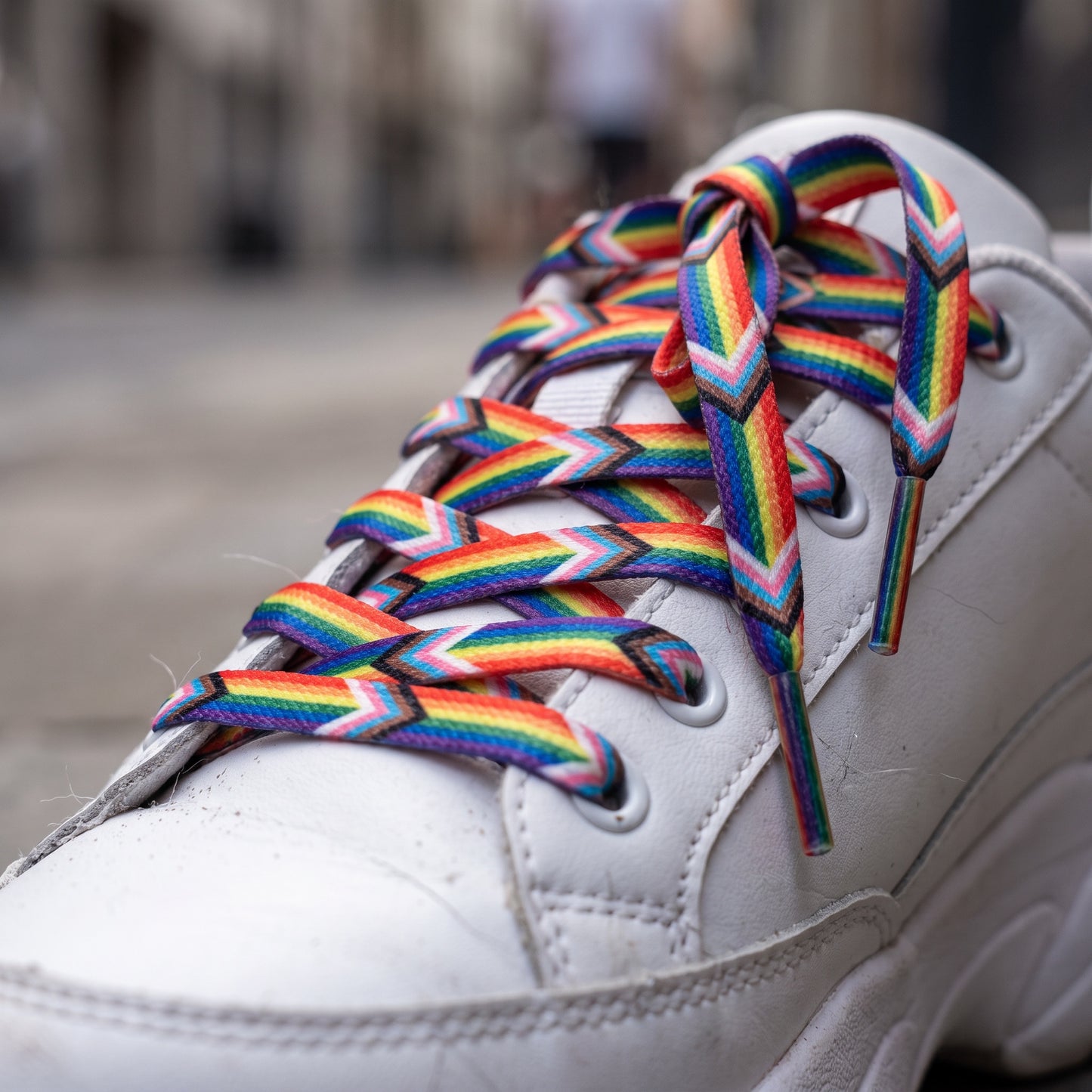 A close-up of a white sneaker laced with RainBee Designs LLC's Pride Shoelace Pack, tied in a bow. The blurred background hints at an outdoor urban setting.
