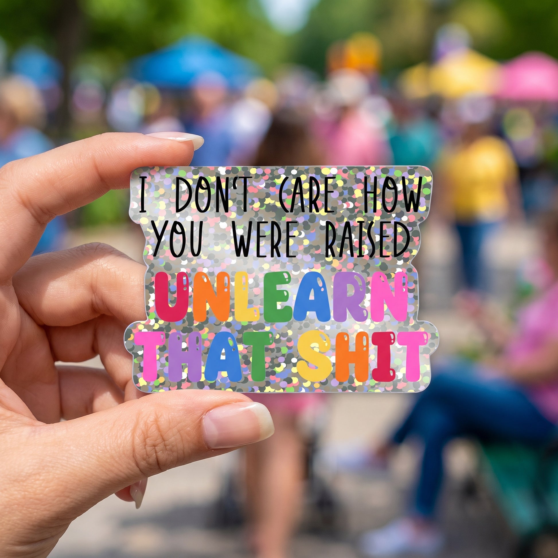 A hand holds a vibrant Unlearn that Shit Glitter Sticker from RainBee Designs LLC, featuring bold lettering that reads, “I don’t care how you were raised. Unlearn that shit,” set against the blurred backdrop of an outdoor event. This eye-catching waterproof vinyl sticker is perfect for making a statement wherever you go.