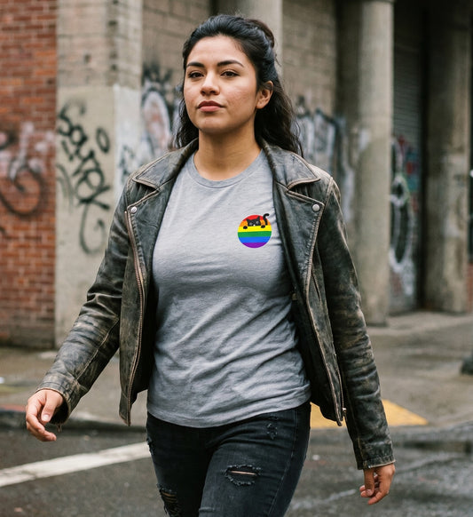 A person wearing the Cat T-Shirt by RainBee Designs LLC—featuring a gray fabric with a rainbow pride heart and black cat graphic—walks along a city street lined with graffiti-covered walls.