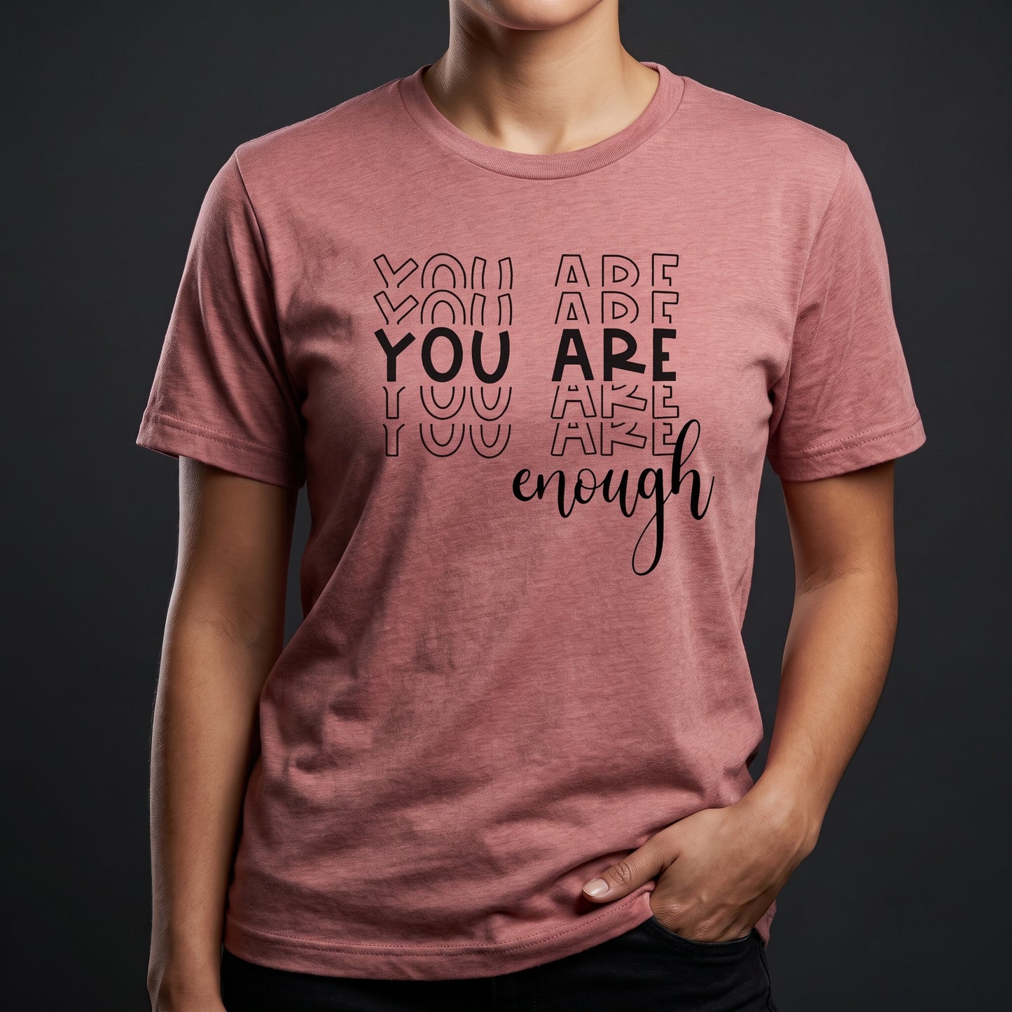 A person wearing a pink RainBee Designs LLC You Are Enough T-Shirt, featuring the words "YOU ARE YOU ARE YOU ARE enough" printed on it, stands against a dark gray background.