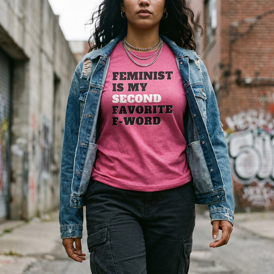 Wearing the “Feminist is My Second Favorite F-Word T-Shirt” by RainBee Designs LLC, a person strides confidently down an urban alley. Layered with a denim jacket and dark cargo pants, this bold fuchsia statement tee—crafted from luxuriously soft tri-blend fabric—seamlessly pairs comfort with empowered attitude.