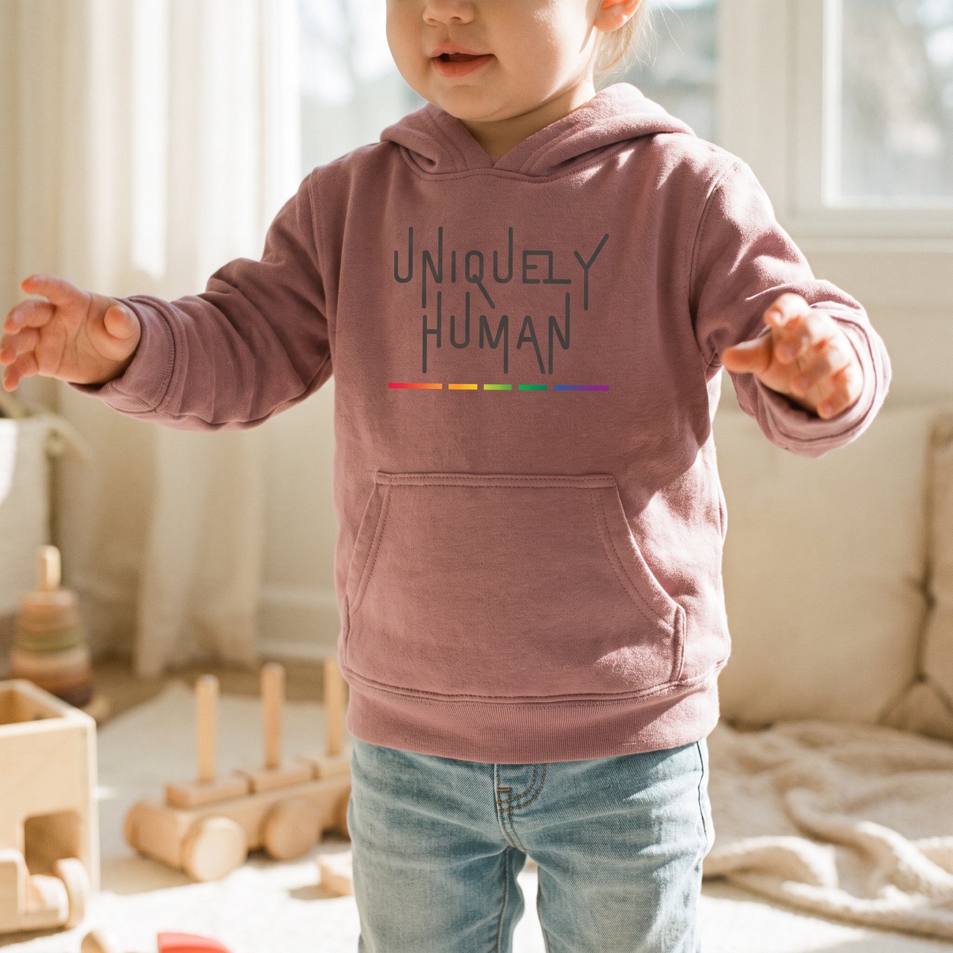 A toddler in the Uniquely Human Toddler Hoodie by RainBee Designs LLC—made from sustainably produced fabric and accented with a rainbow line—stands with arms outstretched in a softly lit, cozy playroom, embodying the spirit of toddler activist apparel.
