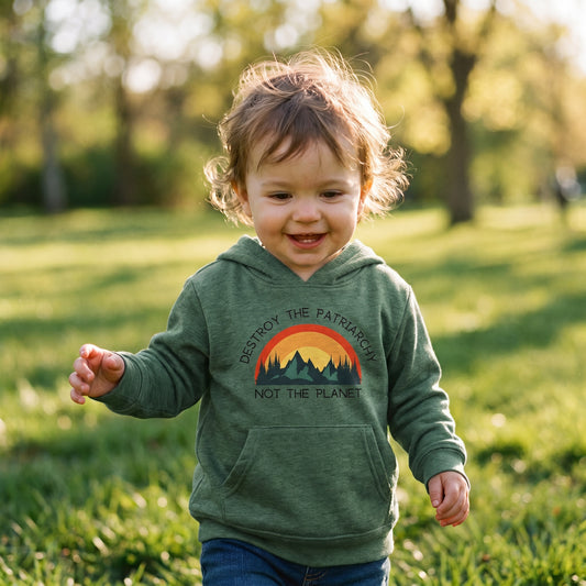 A smiling toddler walks on the grass in a sunlit park, wearing the green Destroy the Patriarchy Toddler Hoodie by RainBee Designs LLC, a standout piece of sustainable kids clothing.