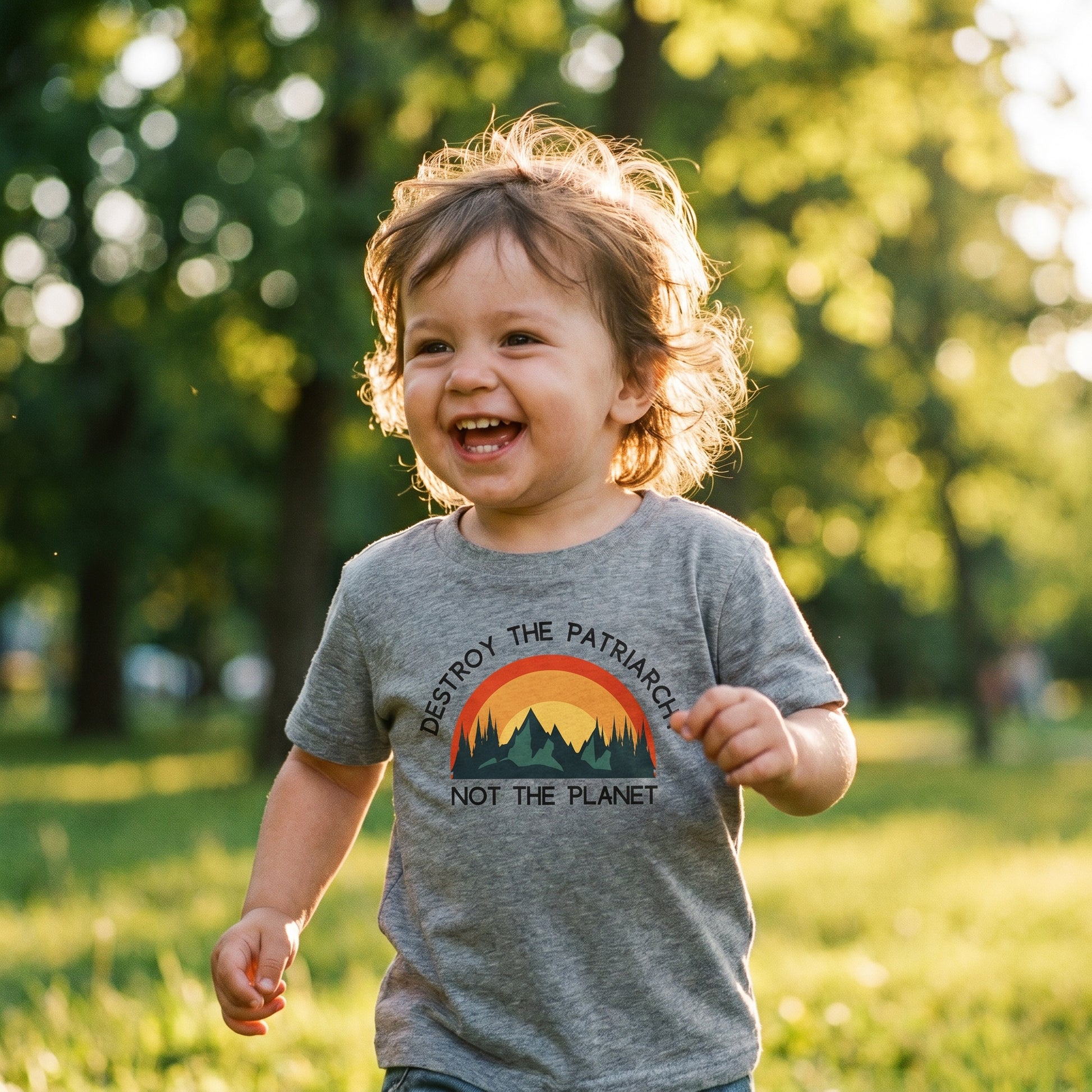 A smiling toddler runs through a sunlit, grassy park, wearing the RainBee Designs LLC Destroy the Patriarchy Toddler T-Shirt—a charcoal heather grey tee featuring a sunset graphic and the words "Destroy the Patriarchy Not the Planet.
