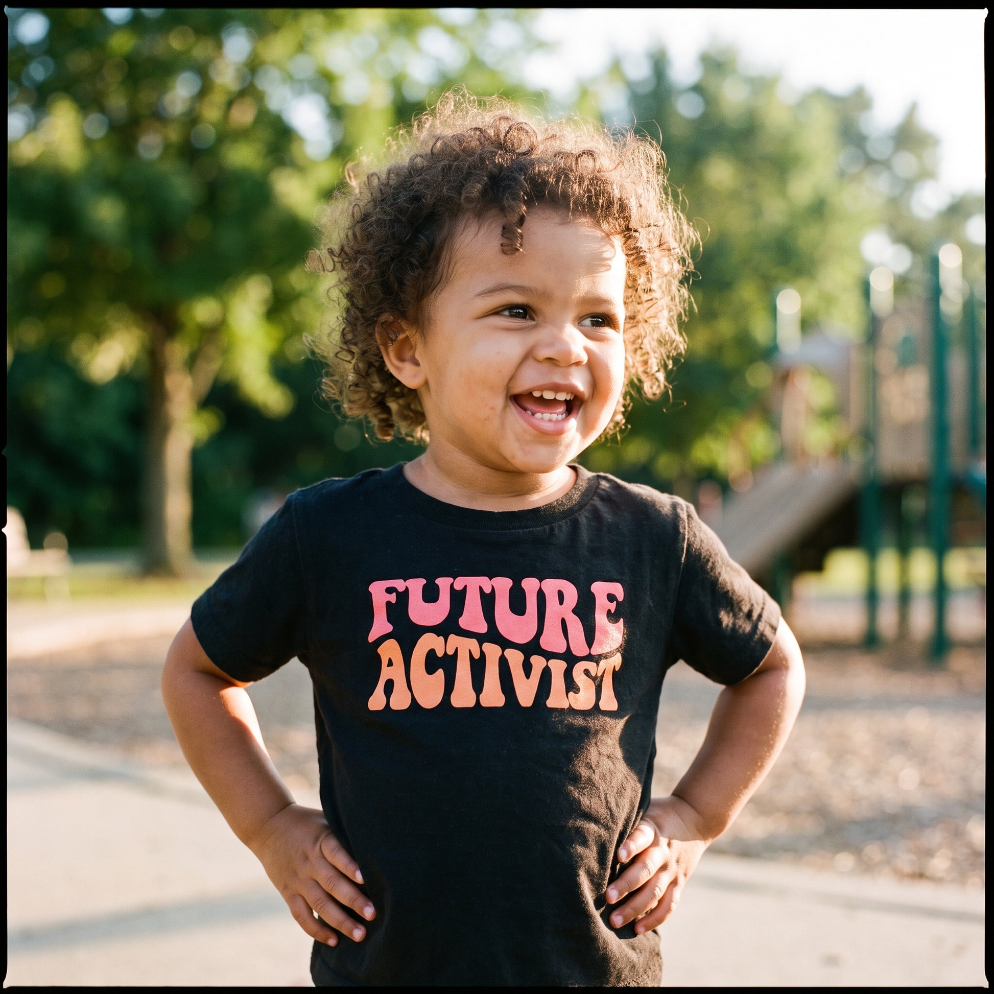 A smiling toddler with curly hair stands outdoors in a park, proudly wearing the RainBee Designs LLC Future Activist Toddler Shirt—one of our standout kids' graphic tees. The black T-shirt features "FUTURE ACTIVIST" printed in bold, colorful letters, perfectly capturing the spirit of children's activist wear. Playground equipment and trees can be seen in the background.