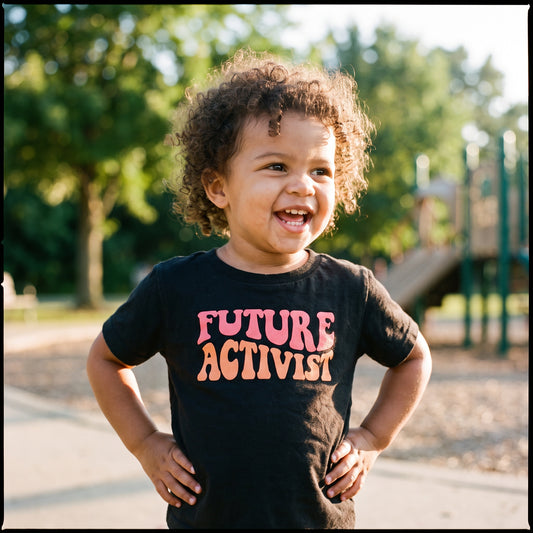A smiling toddler with curly hair stands outdoors in a park, proudly wearing the RainBee Designs LLC Future Activist Toddler Shirt—one of our standout kids' graphic tees. The black T-shirt features "FUTURE ACTIVIST" printed in bold, colorful letters, perfectly capturing the spirit of children's activist wear. Playground equipment and trees can be seen in the background.
