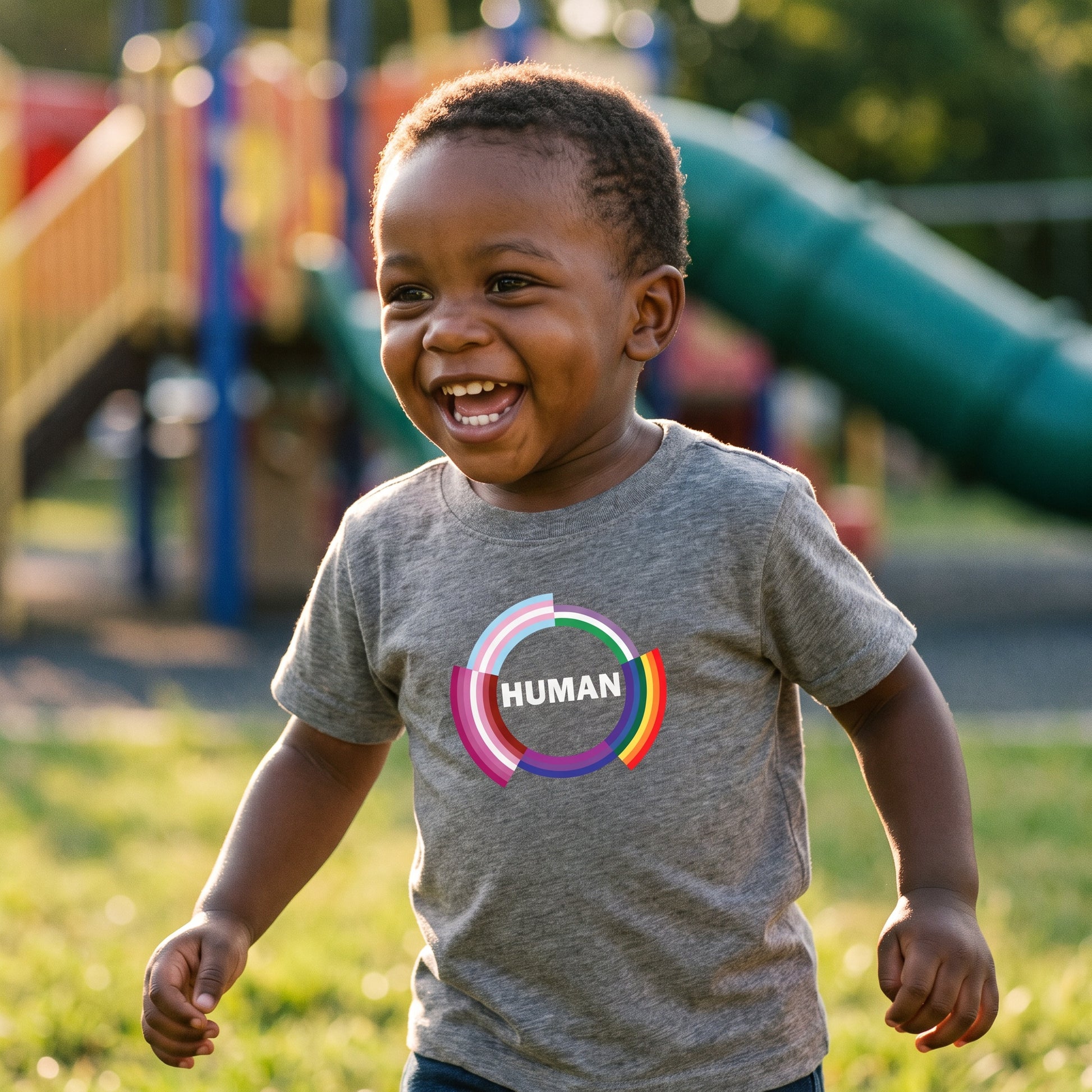 A smiling young child, dressed in the RainBee Designs LLC Human Toddler T-shirt, featuring soft cotton fabric and a bold "Human" print, plays with colorful rings near playground equipment on a sunny day.