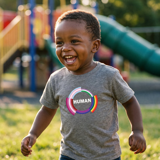 A smiling young child, dressed in the RainBee Designs LLC Human Toddler T-shirt, featuring soft cotton fabric and a bold "Human" print, plays with colorful rings near playground equipment on a sunny day.