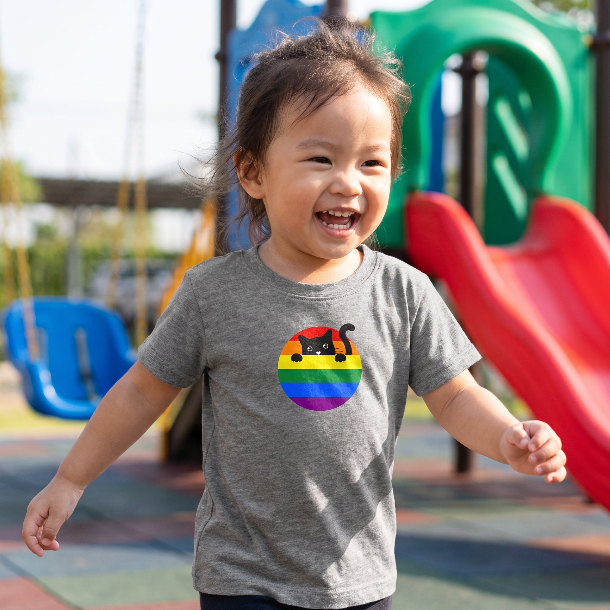 A smiling toddler plays outdoors in front of colorful playground slides and swings, wearing the ultra-soft RainBee Designs LLC Cat Toddler T-shirt featuring a rainbow and black cat design.