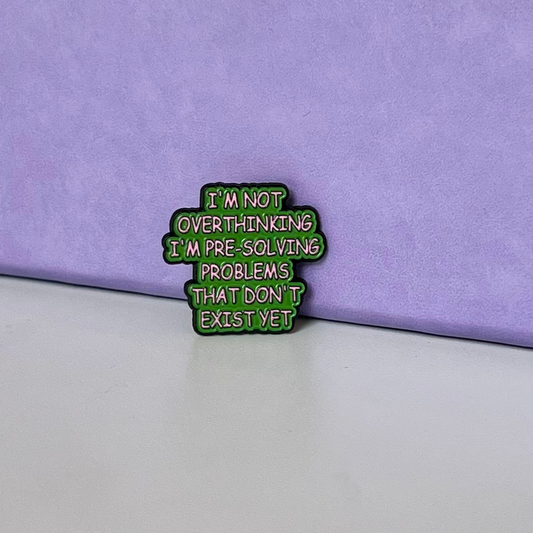 A small enamel pin with the text 'I'M NOT OVERTHINKING I'M PRE-SOLVING PROBLEMS THAT DON'T EXIST YET' in pink lettering on a green background, placed on a light gray surface against a textured purple backdrop.