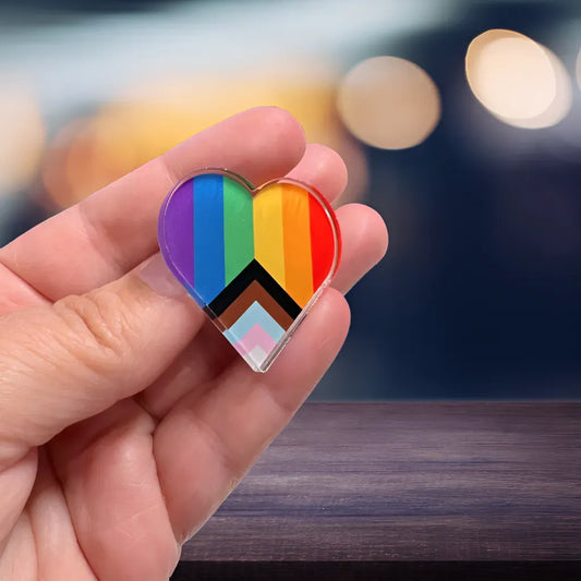 A hand holds the Acrylic Pride Heart Pin by RainBee Designs LLC, featuring Progress Pride flag stripes and chevrons in vibrant acrylic, shining against a softly blurred background.